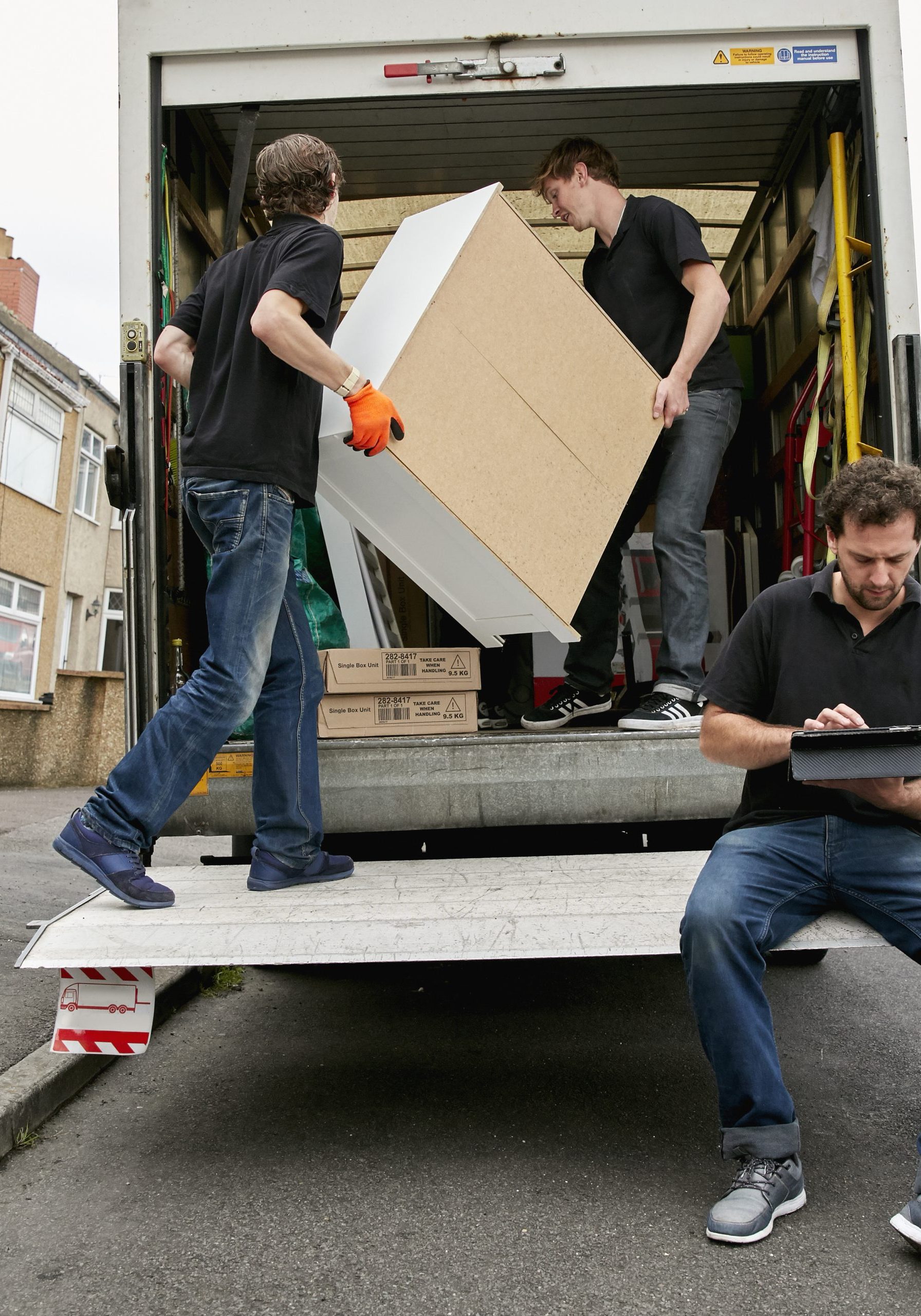 Removals business. A removals company, two men lifting furniture and one seated using a digital tablet.